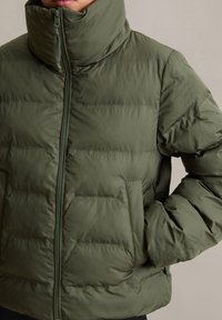 Doudoune vert olive avec col montant, fermeture éclair, texture matelassée et poches latérales. Fabriquée en matière lisse et légère.