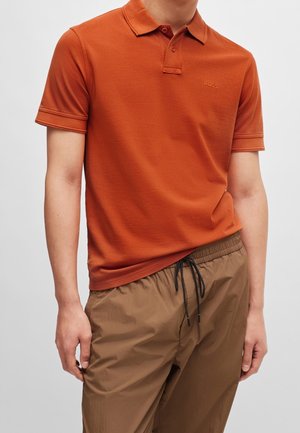 Homme portant un polo à manches courtes orange brûlé et un pantalon marron clair à cordon, debout devant un fond clair uni.