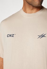 Beež puuvillane t-särk ümarate kaelustega, millel on väike tumehall "CNZ" logo ja dünaamiline graafiline kujundus vasakul küljel.