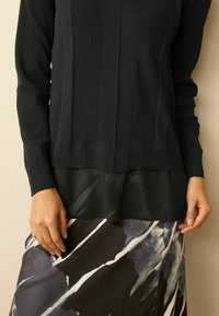 Schwarzer, gerippter Pullover mit einem schichtigen, durchsichtigen schwarzen Saum. Gemusterter, verlängerten schwarz-weißer Rock mit abstrakten Pinselstrich-Details.