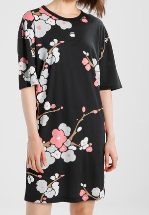 Femme portant une robe noire oversize avec des motifs floraux roses et blancs et des manches courtes, debout devant un fond uni.