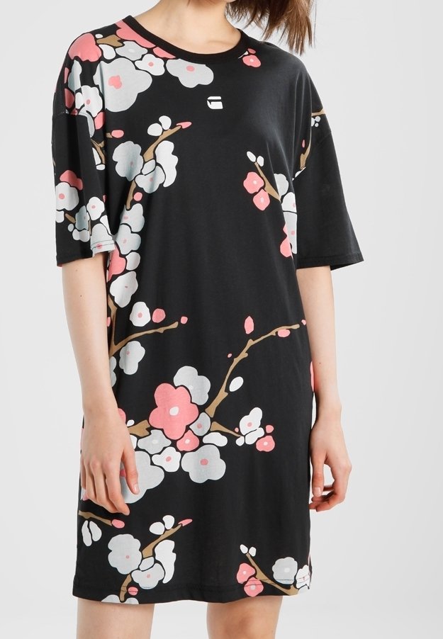 Femme portant une robe noire oversize avec des motifs floraux roses et blancs et des manches courtes, debout devant un fond uni.