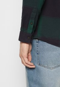 Main gauche reposant à côté de la poche d'un jean en denim bleu clair, portant une chemise à manches longues à carreaux verts foncé et noirs avec un poignet boutonné.