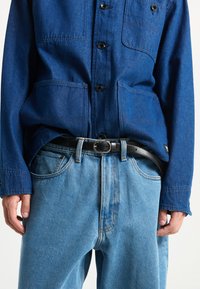 Blaues Denim-Hemd mit Knopfleiste und zwei Brusttaschen, kombiniert mit hellblauen Jeans und einem schwarzen Gürtel.