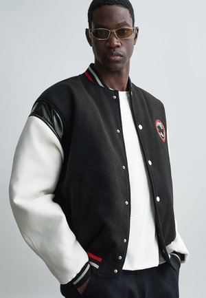 Black and white varsity jacket with leather accents, snap buttons, red and white striped cuffs, and a circular logo patch on the chest.