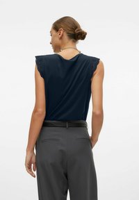 Navy mouwloze top met kanten rand op de schouders, gecombineerd met grijze trousers. Gladde stof en een comfortabele pasvorm, met een subtiele halslijn.