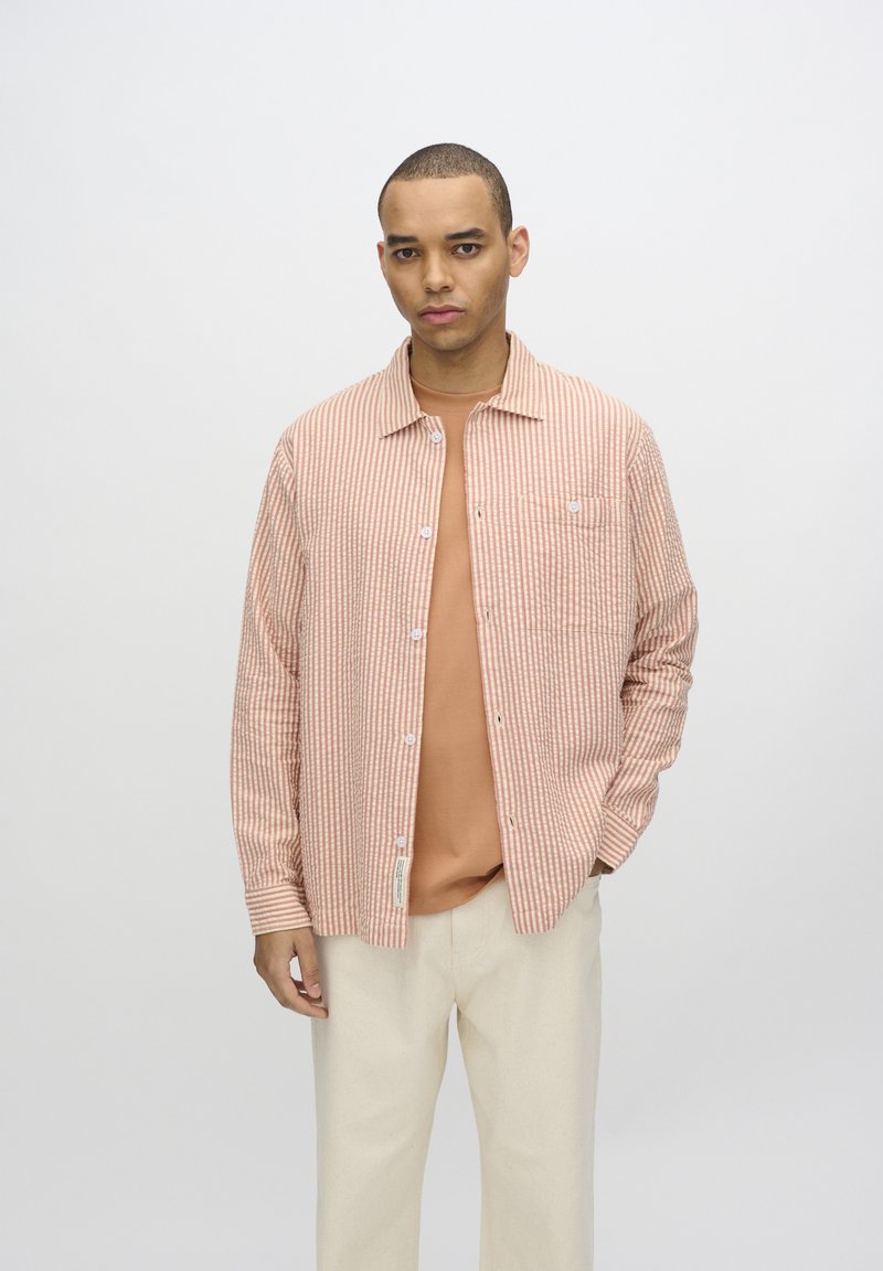 Camisa de botones a rayas en tonos melocotón claro y crema, combinada con una camiseta beige. Textura suave, ajuste relajado y detalle de bolsillo en el pecho.