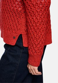 Roter Strickpullover mit einem Lochmuster, gerippten Bündchen, Seitenschlitzen und einem kleinen Metall-Logodetail am Saum. Kombiniert mit dunklem Denim.