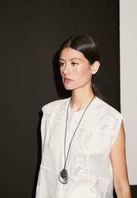 White, short-sleeve blouse with intricate cut-out patterns. A dark pendant necklace hangs from a black cord. Background is dark.