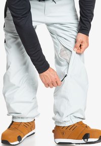 Pantalones de nieve azul claro con ventilación con cremallera, puños elásticos y forro de malla. Se llevan botas impermeables marrones con detalles en negro.