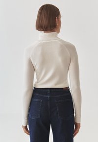 Maglione a collo alto color crema con colletto a coste; design aderente con maniche lunghe, abbinato a jeans in denim scuro con tasche posteriori.