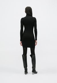 Czarna długorękawowa sukienka przylegająca z wysokim kołnierzem, wykonana z gładkiej tkaniny, zestawiona z czarnymi skórzanymi kozakami do kolan z zamkiem błyskawicznym.