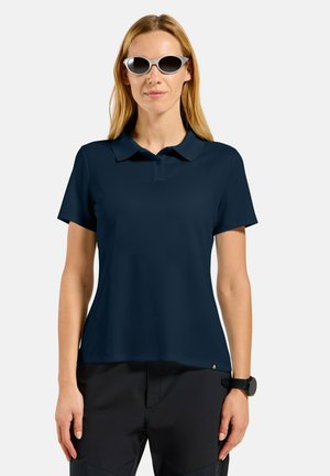 Femme portant un polo bleu marine foncé, un pantalon noir, des lunettes de soleil ovales à monture blanche, et une montre-bracelet noire, debout devant un fond uni.