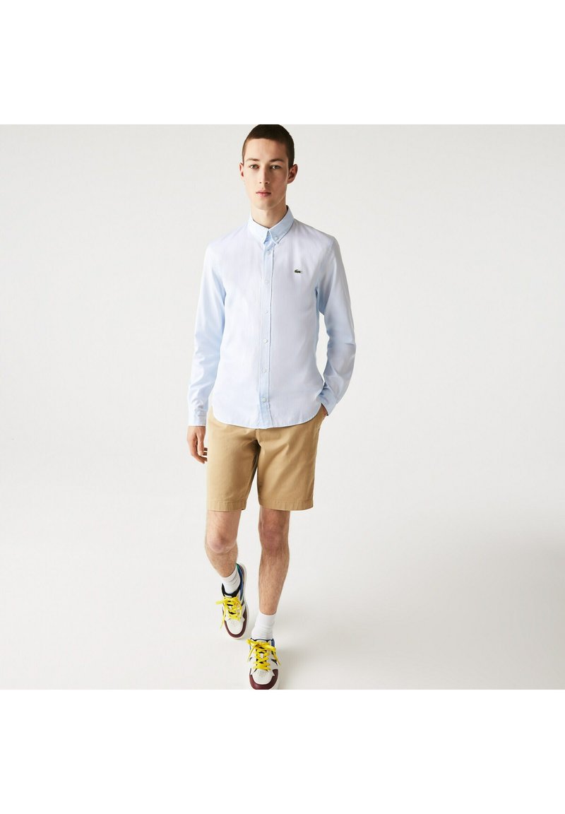 Light blue long-sleeve shirt with a button-down collar, paired with beige shorts. Features a small logo on the chest and colorful sneakers.