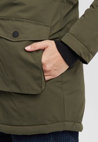 Olive green padded jacket featuring a side pocket and ribbed cuffs. The fabric has a smooth texture with subtle horizontal lines.