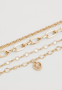 Quatro pulseiras douradas exibidas horizontalmente, apresentando vários estilos de corrente, uma com pedras redondas transparentes e outra com um encantamento em forma de coração.