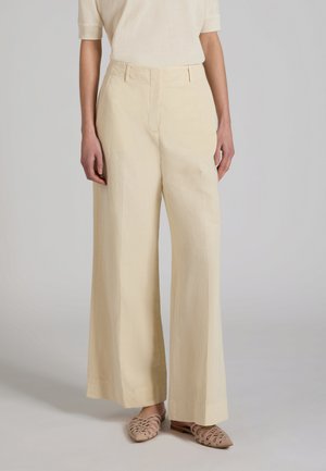 Persona che indossa pantaloni beige a vita alta e gamba larga abbinati a scarpe basse beige intrecciate senza lacci su uno sfondo neutro.