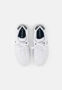 Białe skórzane sneakersy Lacoste z sznurowanym zapięciem. Posiadają czarną wkładkę, zielone logo krokodyla oraz szare akcenty na pięcie.