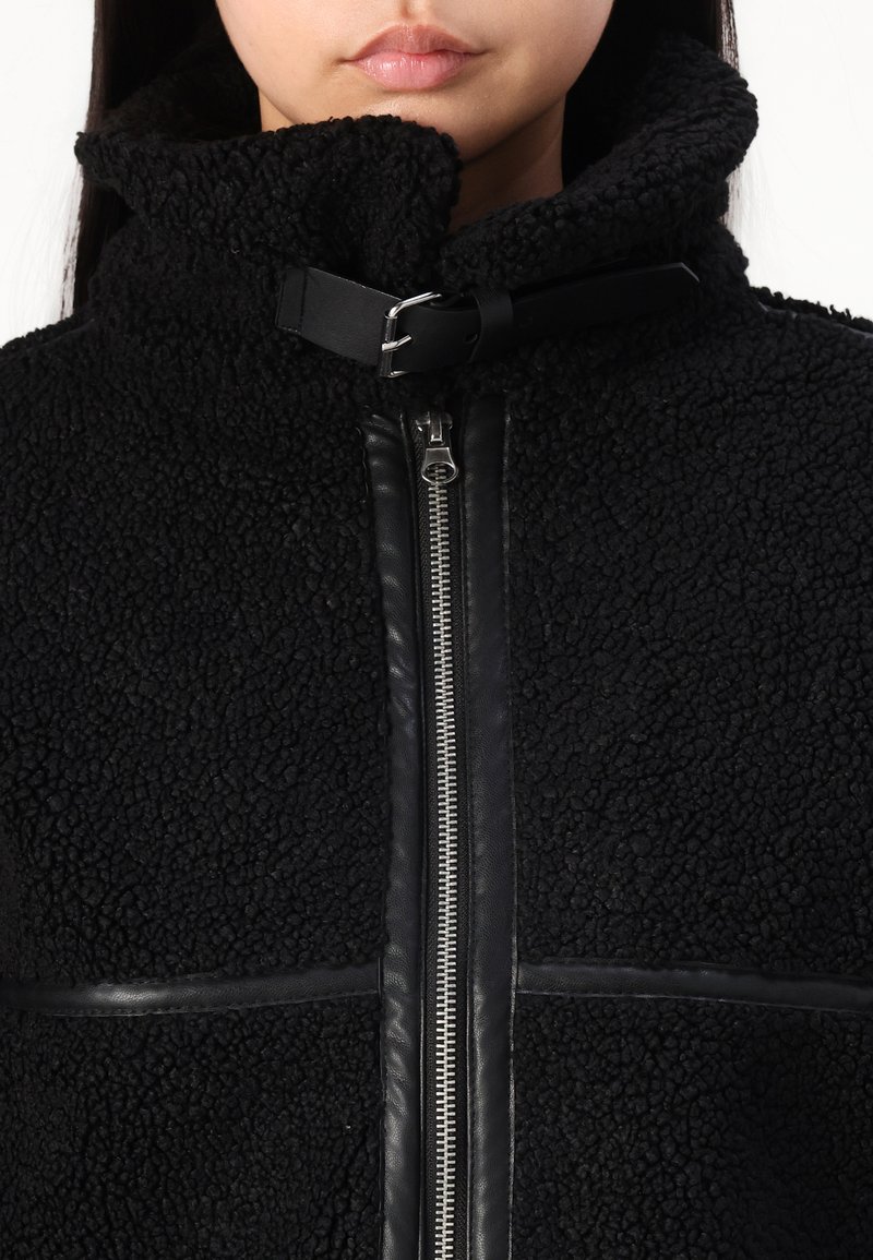 Giacca in pile nera con colletto alto, chiusura zip e dettagli in pelle con finitura testurizzata. Design aderente e casual.