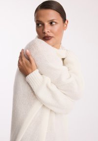 Biały, oversize'owy sweter wykonany z miękkiego, puszystego materiału. Posiada wysoki dekolt i ściągacze na rękawach. Model przytula sweter, eksponując jego fakturę.
