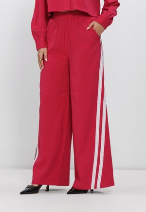 Vrouw die rode wijde broek met witte zijkstrepen draagt, gecombineerd met zwarte pumps met puntige neus en een bijpassend rood cropped topje.