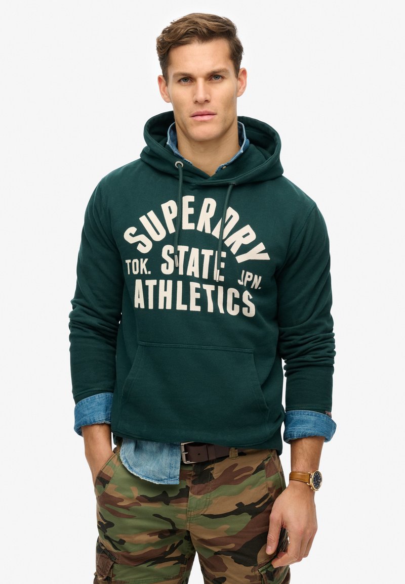 Sudadera verde con capucha y estampado blanco "SUPERDRY ATHLETICS", llevada sobre una camisa de mezclilla azul clara. Combinada con pantalones de camuflaje y un reloj marrón.