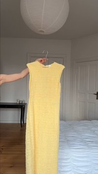 Sleeveless yellow textured dress held on hanger in a minimalist bedroom with white walls, wooden floor, bed, and paper lantern ceiling light.