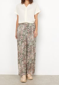 Blouse blanche à manches courtes associée à un pantalon ample à motifs paisley verts, roses et beiges. Portée avec des chaussures beige épaisses.