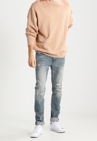 Felpa beige con vestibilità comoda, abbinata a jeans chiari strappati e sneakers bianche. Texture morbida, con risvolti sui jeans.