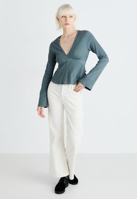 Topp i teal med flagrande ärmar, inslag av en omslagssdesign, strukturvävt tyg och V-ringning. Kombinerad med vita höga byxor och svarta plattformsskor.