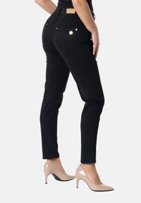 Nainen, joka pukeutuu mustiin slim-fit-farkkuihin ja beigenvärisiin kiiltonahkaisiin korkeakorkoisiin kenkiin, seisomassa sivuttain valkoista taustaa vasten.