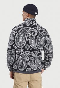 Giacca in pile a motivi paisley in bianco e nero con colletto alto, tessuto testurizzato e polsini elastici, indossata sopra una maglietta grigia.