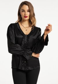Sort bluse med V-hals, struktureret dyreprint, rynket forpanel og lange puffærmer. Knaplukning fortil med let glans.