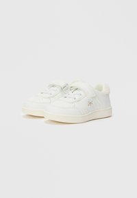 Une paire de baskets blanches pour enfants avec des bandes auto-agrippantes, des détails en dentelle et une semelle beige, arborant un petit logo sur le côté.