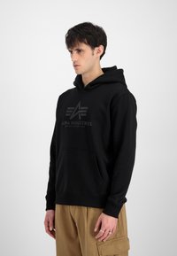 Zwarte hoodie met een voorzak, verstelbare capuchon en reliëflogo. Zacht stof met ribgebreide boorden en zoom, gestyled met kaki broek.
