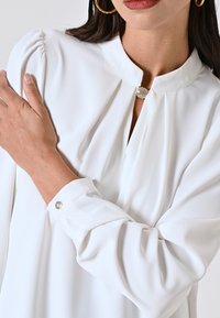 Camicia bianca con colletto alto e design plissettato, caratterizzata da un bottone decorativo al collo e dettagli sui polsini. Tessuto liscio e leggero.