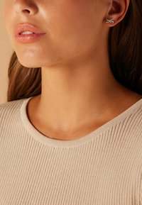 Silberne Steckerohrringe in Form einer Schleife, getragen von einer Person mit heller Haut. Das Oberteil besteht aus geripptem, beigem Stoff mit einem runden Ausschnitt.