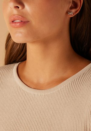Boucles d'oreilles en argent en forme de nœud, portées par une personne à la peau claire. Le haut est en tissu beige à nervures avec un col rond.