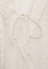 Tissu blanc texturé avec un col en V orné d'un nœud noué et d'un bordure douce à volants le long des bords du col.