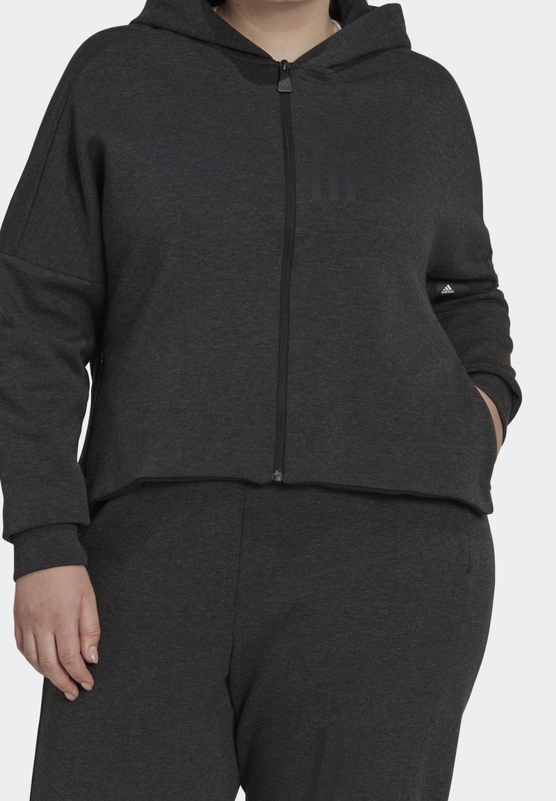 Personne portant une veste à capuche zippée Adidas gris foncé et un pantalon assorti, debout avec une main dans la poche sur un fond clair.