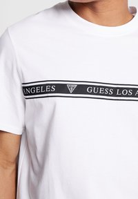 Bílá bavlněná tričko má černý grafický pruh s textem "GUESS LOS ANGELES" a trojúhelníkovým logem uprostřed. Krátké rukávy.