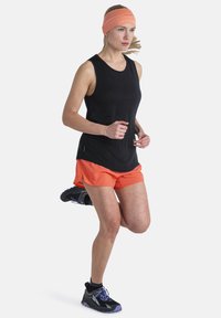 Camiseta sin mangas negra, pantalones cortos de running naranjas y una cinta para la cabeza rosa. El corredor lleva zapatillas deportivas negras con acentos morados.