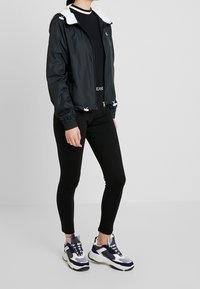 Woman wearing black zip-up hooded jacket, black leggings, and white and navy chunky sneakers standing against plain background.
