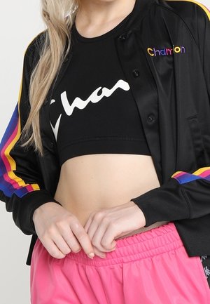 Femme portant un crop top noir et une veste à rayures multicolores, associée à un pantalon rose à taille élastique, les mains reposant près de la taille.