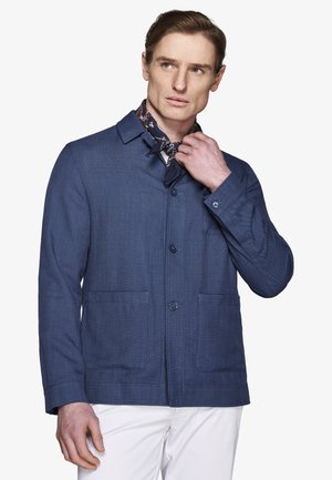 Mężczyzna w niebieskiej zapinanej na guziki kurtce, białych spodniach i kwiecistym szaliku na szyi, poprawiający szalik i patrzący w bok na białym tle.