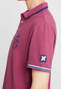 Burgundy pikétröja med navyfärgad krage, broderad logotyp och randiga detaljer på ärmarna. Slät tygtextur, standardpassform.