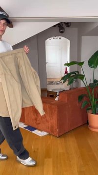 Bež jakna oversized s dugim rukavima, prednjim džepovima i detaljima dugmadi, koju drži osoba u ležernoj odjeći. Vidljiv drveni pod i biljka.