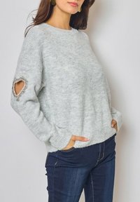 Pull en laine gris avec ourlet et poignets côtelés, présentant une découpe en forme de cœur sur la manche ornée de perles, associé à un jean foncé.