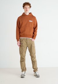Brązowy hoodie z przednią kieszenią, w połączeniu z beżowymi spodniami cargo z wieloma kieszeniami. Model nosi czarno-białe trampki. Minimalne tło.