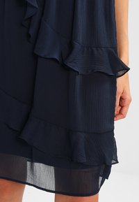 Robe texturée bleu marine avec des détails de volants superposés près de l'ourlet et une main visible reposant à côté de la jambe.